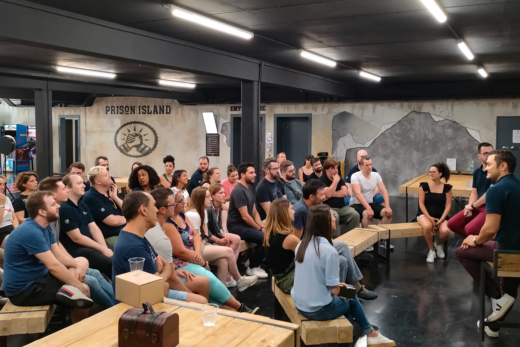 Un groupe d'une quarantaine de collègues assis sur des bancs écoute le briefing devant l'emblème Prison Island